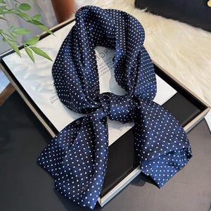 Può includere: Una sciarpa blu navy con pois bianchi. La sciarpa è annodata, con le estremità che pendono. La sciarpa è realizzata in un materiale setoso e probabilmente viene indossata come accessorio di moda.