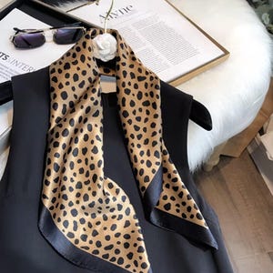 May include: A silk scarf with a tan leopard print and black border is draped over a black garment. The scarf is hanging on a hanger with a white flower accent. Sunglasses and a framed document are in the background.