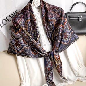 May include: A patterned silk scarf in shades of navy, burgundy, and gold, tied in a knot. The scarf is draped over a white blouse. A black handbag and a magazine with the word "LOEWE" are in the background.