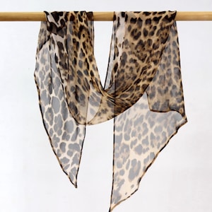 Può includere: Sciarpa trasparente con stampa leopardata. Il tessuto leggero è drappeggiato su un'asta di legno, mostrando un motivo di macchie marroni e nere su sfondo beige. Un accessorio di moda classico e senza tempo.