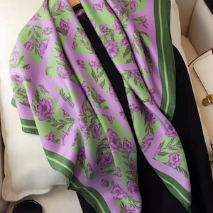 May include: A patterned silk scarf. The scarf displays a repeating design of purple flowers and green leaves on a light green and lavender backdrop. It has a dark green border. The scarf is draped over a hanger, with a pair of sunglasses visible below.