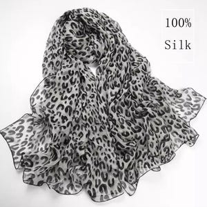 Op de afbeelding: Een lichte zijden sjaal met een zwart-grijs luipaardpatroon. De sjaal is gedrapeerd en toont de vloeiende stof en delicate randen. De tekst "100% Silk" is zichtbaar.