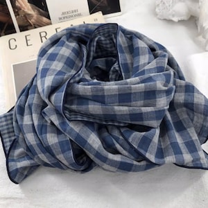 May include: A blue and grey checked scarf, folded and draped, with a dark blue border. The scarf is made of a soft, lightweight fabric. The scarf is lying on top of a book with the word "CER" visible.