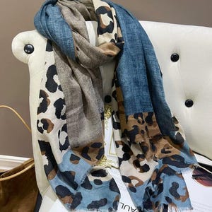 May include: A rectangular scarf featuring a patchwork design of blue denim-textured fabric and beige fabric with a black and brown leopard print pattern. The scarf has frayed edges and is draped in folds, showcasing the contrasting textures and colours.