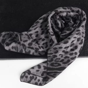 May include: A grey and black leopard print scarf, draped and folded. The scarf's pattern features dark spots on a lighter grey background. The fabric appears soft and lightweight, suitable for various styling options.