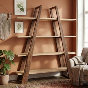 May include: A wooden bookshelf with a leaning ladder design. The frame is dark brown, and the shelves are light brown. The bookshelf has four shelves and is placed against a warm-toned wall. Decorative items and a plant are visible.