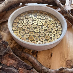 May include: A circular concrete tray filled with clear resin containing numerous brass shotgun shell bases. Each metallic base features the text Fiocchi Italy 12 embedded in the resin, creating a uniform pattern within the light grey concrete rim.