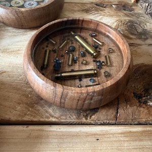 May include: A round wooden bowl featuring a clear resin inlay on the interior base. Embedded within the resin are various brass bullet casings and small metallic lead pellets, creating a textured display of ammunition components.