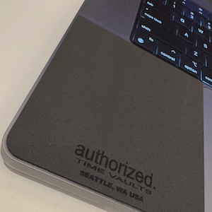 Puede incluir: Un ordenador portátil gris oscuro con un teclado negro y el texto "authorized. TIME VAULTS SEATTLE, WA USA" impreso en la superficie. El portátil está abierto, mostrando el teclado y una parte de la pantalla.