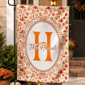 May include: A rectangular garden flag featuring a pattern of orange, yellow, and red autumn maple leaves. In the center, a white oval frame with an orange border contains a large orange letter H and the cursive text The Hazel in brown.