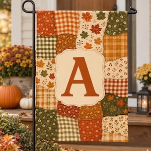 May include: A rectangular garden flag featuring a patchwork design of autumn-themed patterns including orange and green gingham, maple leaves, and small flowers. A large, centered cream-colored scalloped plaque displays the capital letter A.