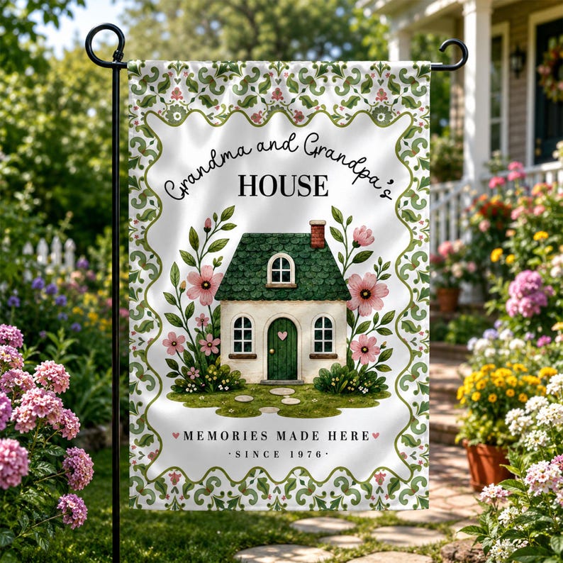 May include: A decorative garden flag featuring a whimsical illustration of a house with a green roof and pink flowers. The flag reads "Grandma and Grandpa's HOUSE" and "MEMORIES MADE HERE SINCE 1976" in a decorative font. The flag is white with a green and pink floral border.