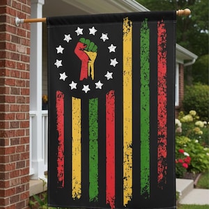May include: Black flag with vertical stripes in red, yellow, and green. A circle of white stars surrounds a raised fist in red, yellow, and green. The flag is hanging on a wooden pole outside a brick house.
