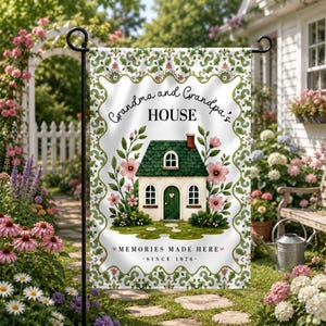 May include: A white garden flag with a green and pink floral design. The flag features a small house with a green roof and the words "Grandma and Grandpa's HOUSE". Text at the bottom reads "MEMORIES MADE HERE SINCE 1976".