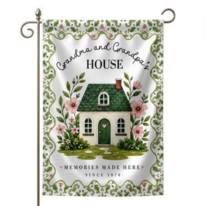 May include: A decorative garden flag featuring a watercolor illustration of a house with a green roof and door, surrounded by pink flowers and greenery. The flag reads "Grandma and Grandpa's HOUSE" and "MEMORIES MADE HERE - SINCE 1976."