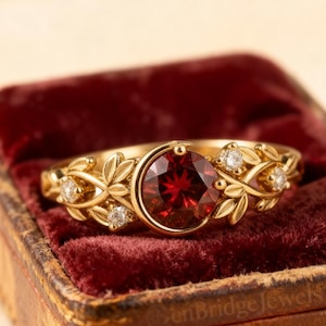May include: A gold ring featuring a large, round, red gemstone, accented with smaller white stones and leaf motifs. The ring is presented in a vintage jewellery box lined with red velvet.