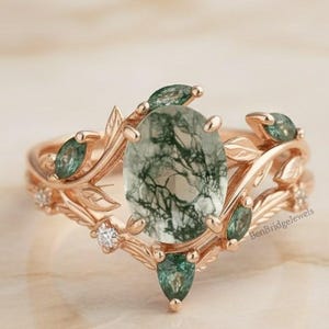 May include: Rose gold engagement ring set with an oval moss agate center stone and leaf and vine detailing. The ring is accented with small green gemstones and tiny diamonds. The set includes a matching band.