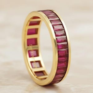 May include: A gold band ring with a continuous row of rectangular, deep red gemstones. The ring's setting is a polished gold, and the gemstones are flush with the band's surface. The ring is a full eternity style.