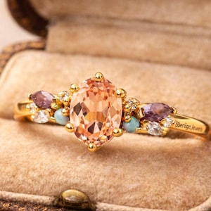 May include: A gold ring with a large peach-colored oval gemstone as the centerpiece. The ring is adorned with smaller purple, turquoise, and clear gemstones. The ring is inside a brown jewelry box with a velvet interior. The ring is engraved with the text "Ebenige Jewels".