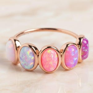 May include: A rose gold ring featuring five oval-shaped opals in shades of pink and purple. The opals are set in individual bezels, creating a delicate and feminine design. The ring is displayed on a marble surface.