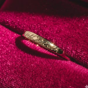 Könnte beinhalten: Ein goldener Ring mit der Inschrift "Till Death" auf dem Band. Der Ring ist vor einem tiefroten Samthintergrund platziert, was eine luxuriöse und romantische Ästhetik erzeugt.