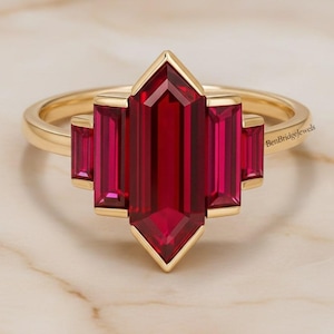 May include: A gold ring with a central, rectangular, ruby-red gemstone, with two smaller rectangular stones on either side. The ring has a geometric design and is set against a marble background. The brand name "BenBridgeJewels" is visible.