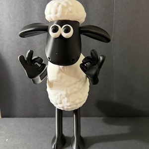 以下が含まれることがあります： アニメシリーズのキャラクター、ひつじのショーンの3Dプリントフィギュア。 羊は白い体、黒い顔、耳、脚を持ち、両手で「OK」のハンドジェスチャーをしています。 フィギュアは灰色の表面に立っています。