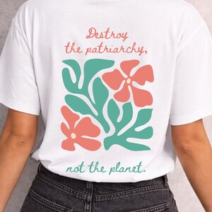Könnte beinhalten: Weißes T-Shirt mit dem Text "Destroy the patriarchy, not the planet." Das Design zeigt korallenfarbene Blumen und türkisfarbene Blätter. Das Shirt besteht aus einem weichen Material.