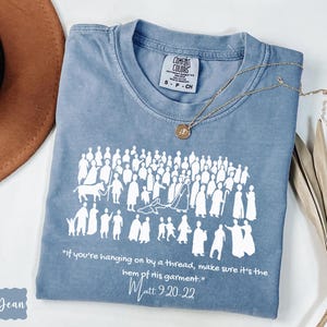 May include: A blue Comfort Colors t-shirt with a white graphic of people and text from Matthew 9:20-22. A gold necklace and a brown hat are next to the shirt. The shirt has the words "Blue Jean" on the bottom left.