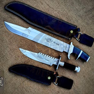 May include: Two silver-toned steel knives with black handles and matching black leather sheaths. The larger knife is marked "LIFE SUPPORT PREDATOR" and the smaller knife is marked "LIFE SUPPORT EXTRA I". Both blades feature a metallic finish.