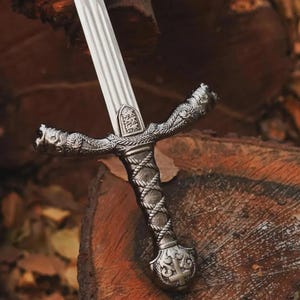 May include: A detailed close-up of a silver sword with an ornate handle and crossguard. The blade is ridged, and the handle features a woven design. The crossguard has decorative elements, and the sword rests on a wooden surface.