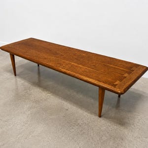 May include: A rectangular wooden coffee table with a medium brown finish, featuring rounded corners and four tapered legs. The tabletop displays a visible wood grain pattern and distinct dovetail joinery details at the corners of the surface.