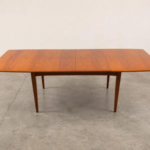 May include: A rectangular wooden dining table with a warm brown finish and visible wood grain. The table features a smooth, flat surface with a central seam indicating an extendable design, supported by four tapered legs that angle outward.