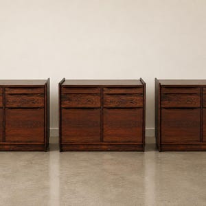 May include: Three matching dark brown wooden cabinets, each featuring two upper drawers with horizontal pulls and two lower cabinet doors. The wood grain is prominent and dark, with a rectangular frame and a solid base for each unit.
