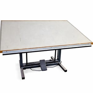 May include: A large, adjustable drafting table with a light-colored, rectangular work surface. The table has a metal frame and a foot pedal for height adjustment. The work surface is approximately 48 inches wide and 36 inches deep.