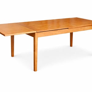 May include: A light brown wooden dining table with an extendable leaf. The table has a rectangular top and four legs. The leaf is extended, increasing the table's surface area. The table is made of wood and has a natural finish.