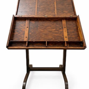 May include: A vintage wooden drafting table with a hinged top and a lower storage area. The table is made of dark-stained wood and has a sturdy, curved-leg base. The top is open, revealing the interior.