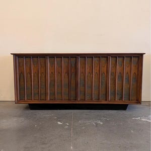 May include: A long, rectangular wooden sideboard featuring four cabinet doors with vertical, arched slats. The wood has a rich brown finish, and the cabinet sits on a recessed black base. The design includes repeating geometric patterns.