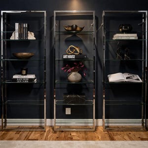 May include: Three modern glass and chrome display shelves against a dark blue wall. Each shelf unit is filled with decorative items like books, bowls, sculptures, and a vase with flowers. The shelves are on a wooden floor.