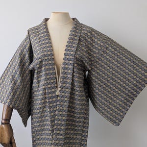 May include: A short, open-front jacket with wide, kimono-style sleeves. The garment features a repeating geometric pattern in shades of dark blue, gold, and white. It has a structured collar and a simple tie closure at the center of the neckline.