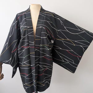 Può includere: Una giacca haori nera caratterizzata da un motivo di sottili linee bianche astratte, intervallate da occasionali accenti rossi, gialli e verdi. Il capo presenta maniche ampie e larghe e un design aperto sul davanti, esposto su un manichino.