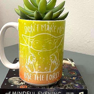 May include: A yellow mug with a white handle, featuring a cartoon character and the phrases "DON'T MAKE ME" and "USE THE FORCE." The mug holds a green succulent plant and rests on a book titled "A Mindful Evening."