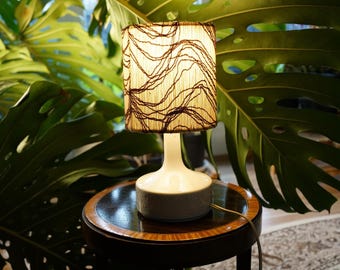 May include: A small, white table lamp with a cylindrical base and a rectangular lampshade. The lampshade is beige with a brown, abstract design. The lamp sits on a dark, round table with a decorative edge, in front of large green leaves.