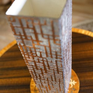 Op de afbeelding: Een hoge, rechthoekige vaas met een geometrisch patroon in bruin- en grijstinten. De vaas heeft een vierkante opening en staat op een decoratieve, ronde onderzetter. Het ontwerp doet denken aan een architecturaal motief.