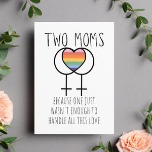 May include: A white greeting card with the text "TWO MOMS" above a symbol of two connected Venus symbols, with a rainbow heart in the center. The text "BECAUSE ONE JUST WASN'T ENOUGH TO HANDLE ALL THIS LOVE" is below the symbol.