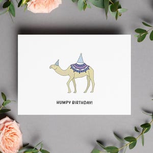May include: A white greeting card with a cartoon camel wearing a party hat and a decorated blanket. The text "HUMPY BIRTHDAY!" is printed below the camel in black. The card is surrounded by pink roses and green leaves.