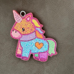 May include: A colorful unicorn-shaped charm with glitter accents. The charm features a pink outline, a gold horn, and a blue body with an orange heart. The mane and tail are yellow and green. A silver ring is attached at the top.