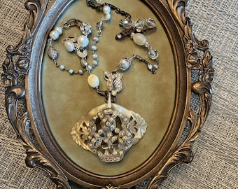 May include: An antique oval-shaped, bronze-colored frame with ornate floral detailing surrounds a green velvet interior. Several pieces of jewelry, including necklaces and a pendant, are displayed within the frame. The jewelry features pearls and clear gemstones.