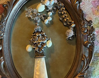 May include: An antique silver-toned serving utensil with a mother-of-pearl handle, displayed in an ornate oval frame. The frame holds the utensil and several pieces of jewelry, including a pearl necklace and a crystal and pearl brooch. The background is a floral pattern.