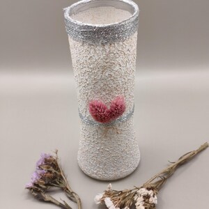 Peut inclure: Un vase blanc texturé avec un bord argenté et une bande argentée au milieu, orné de deux accents floraux roses en forme de cœur. Des fleurs séchées sont placées près du vase. Le vase mesure environ 20 cm de haut.